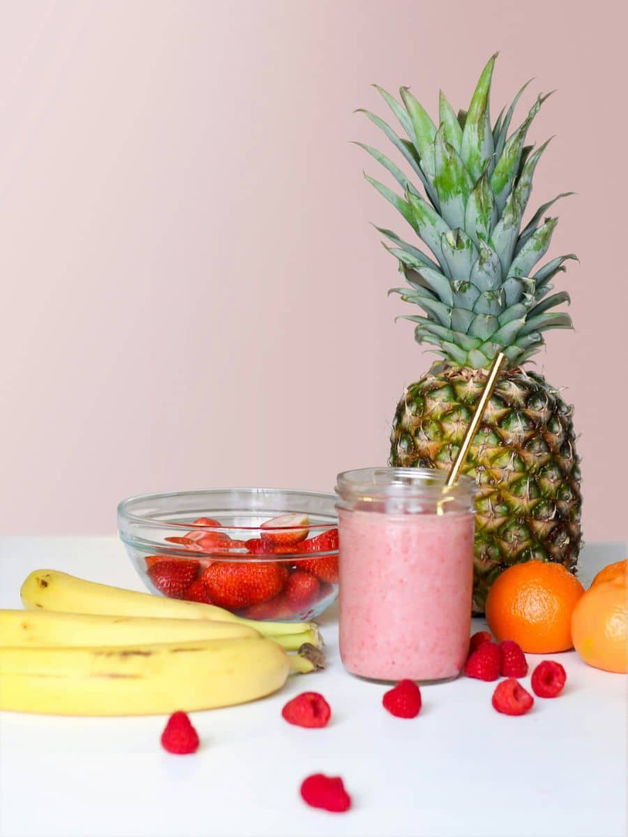 Pink slushie sitting next to a pineapple, bananas, raspberries, and oranges.