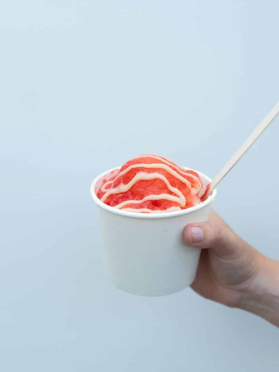 Reddish colored slushie drizzled with something white in a white cup with a spoon sticking out being held by a hand.