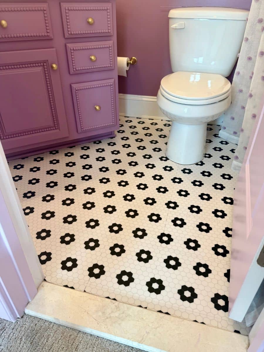 White tile with black flowers on the floor of a bathroom with a purple vanity and white toilet.