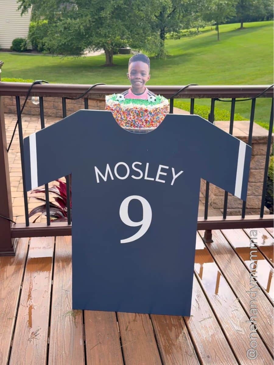 Sports jersey cake stand made out of wood, painted blue, and has the name Mosley and the number 9 on it with a confetti cake and a a boy's head next to soccer balls.