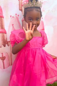 Little girl in a pink dress with a gold crown holding up 4 fingers for her four year old birthday.