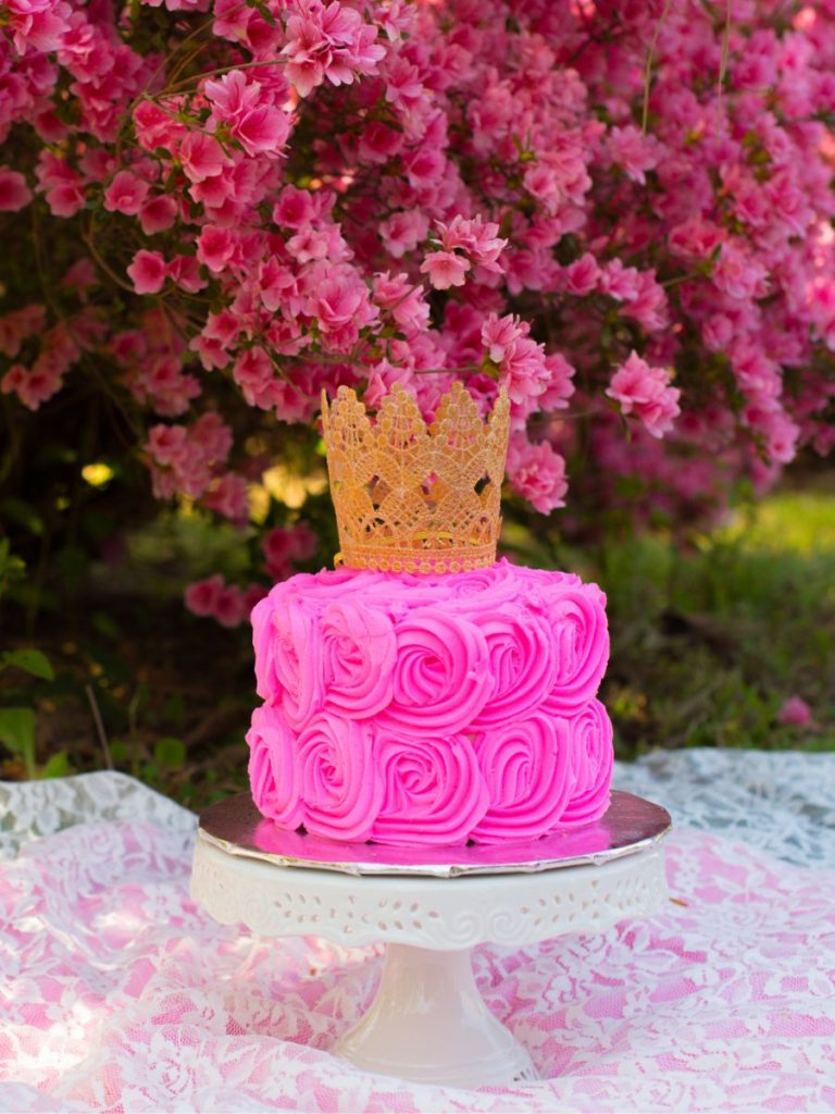 Cake with swirly pink icing topped with a gold crown sitting on a silver and white pedestal platter