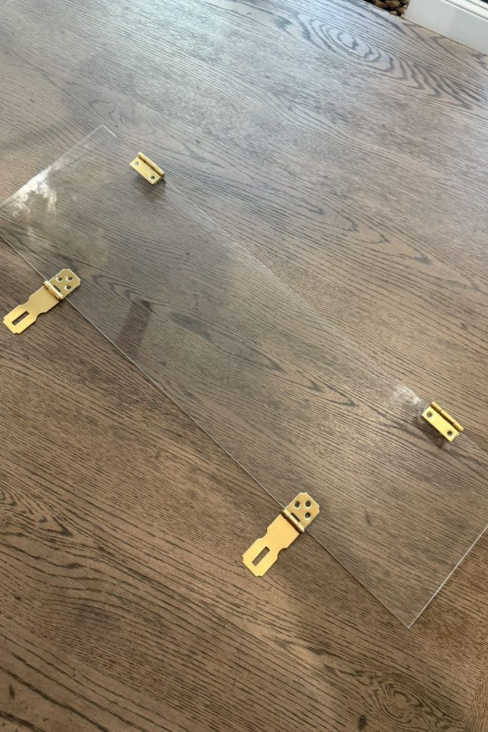 Picture of a piece of plexiglass with gold hinges and gold latches on a dark wood background.