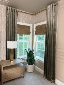 Corner of a room showing a small table with drawers and a lamp on top, two windows with naturally woven shades and elegant velvet curtains, and a green plant.