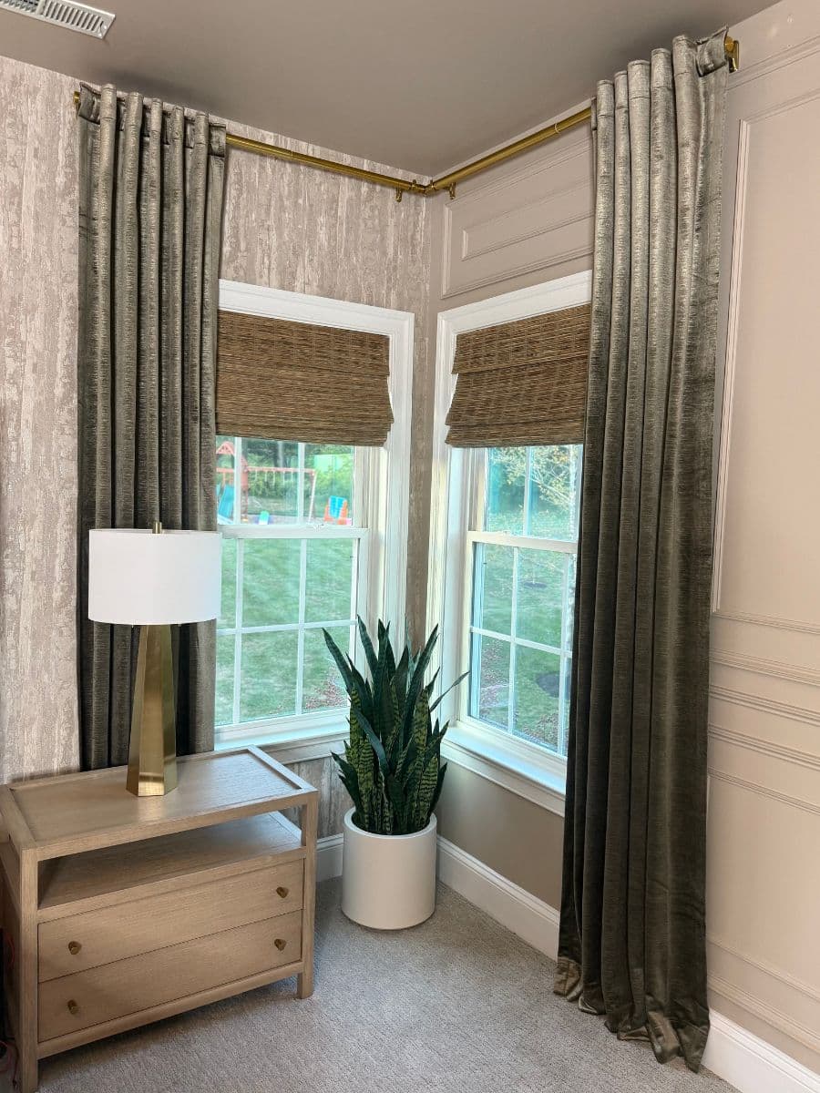 Corner of a room showing a small table with drawers and a lamp on top, two windows with naturally woven shades and elegant velvet curtains, and a green plant.