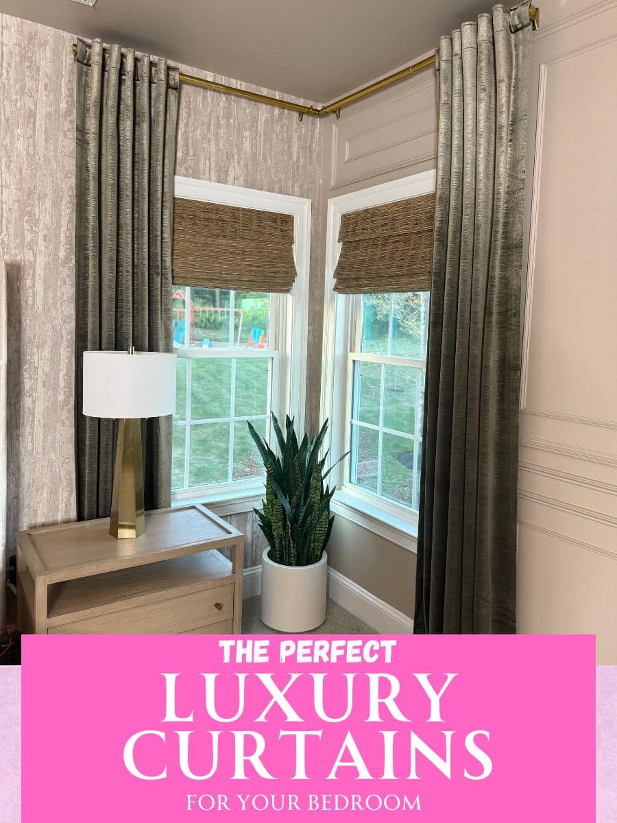 The corner of a room that has a small table with drawers and a lamp on top, two windows with natural brown shades and dark velvety curtains on a gold curtain rod, and a green plant.